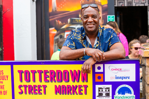 A smiling man in a colourful shirt leans on a sign that says 'Totterdown Street Market' in vibrant writing