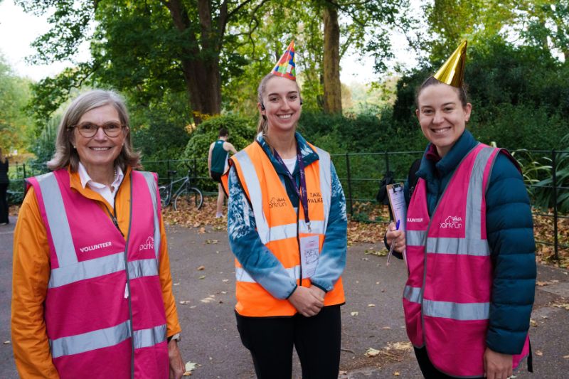 Three people wearing high-visibility vests, two pink marked 'VOLUNTEER' and one orange, stand in a park. Two of them wear festive party hats. Trees and a person near a bicycle are visible in the background.
