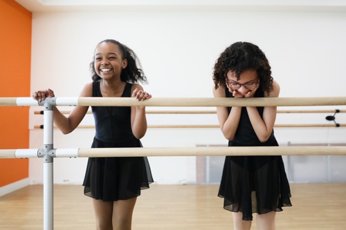 Two young Black ballerinas stand at a dance studio barre laughing and smiling