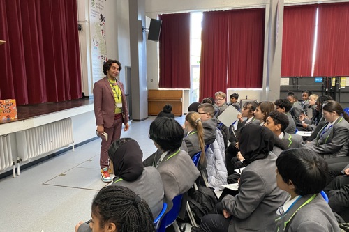 Author Joseph Coelho stands in front of a room of school pupils dressed in grey and green uniforms